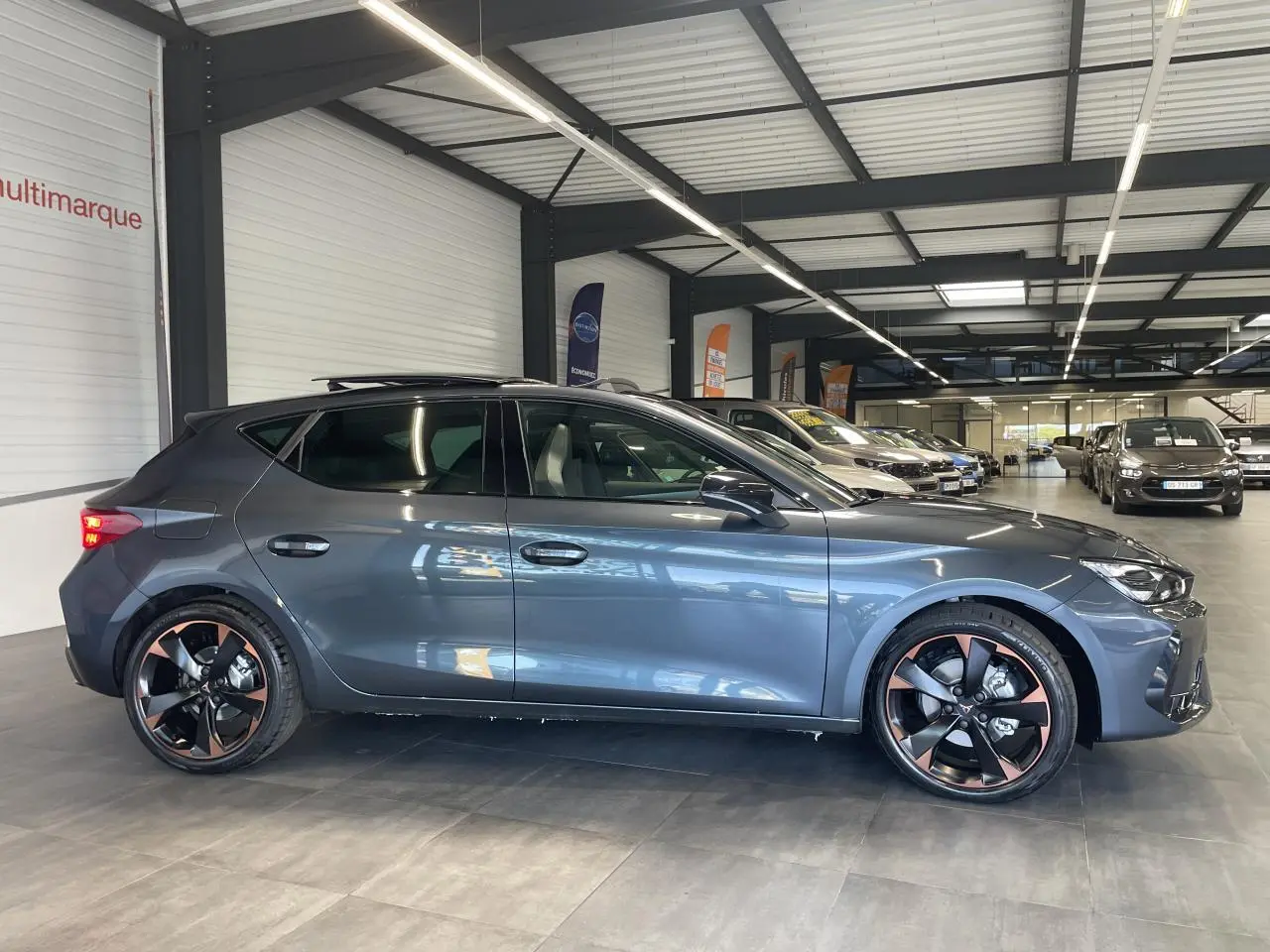 Vue de profil côté gauche d'une CUPRA Leon gris magnétique avec jantes alliage 18'' cuivrées en showroom.