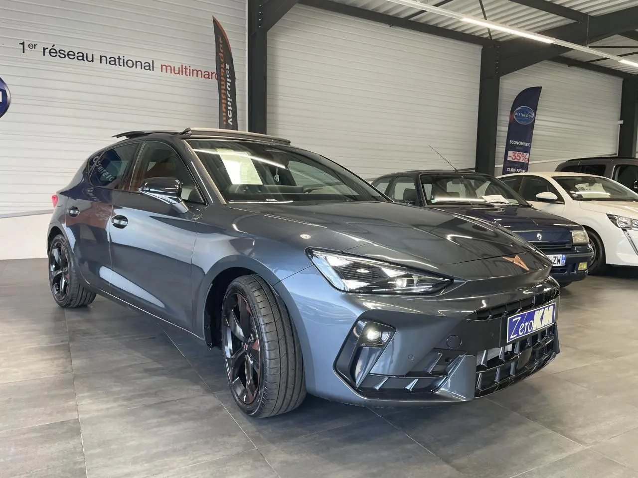 Vue 3/4 avant droite d'une CUPRA Leon gris magnétique avec jantes alliage noires et toit ouvrant panoramique en showroom.