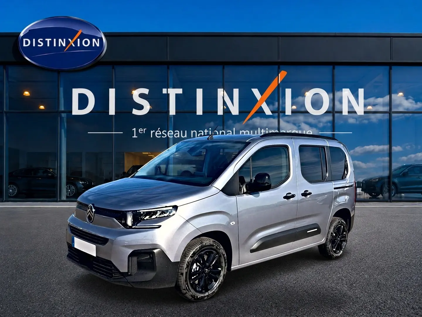 Citroën Berlingo Steel Grey vue 3/4 avant droit, avec jantes noires et barres de toit visibles devant vitrine Distinxion.