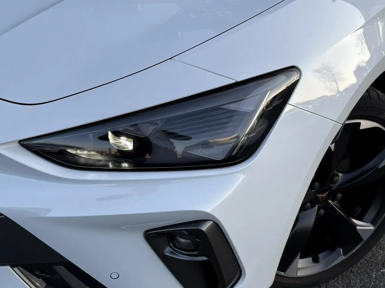 Gros plan sur l'avant droit blanc Neige d'une CUPRA Leon 2025, mettant en valeur le phare Tri-LED et la jante alliage 18 pouces.