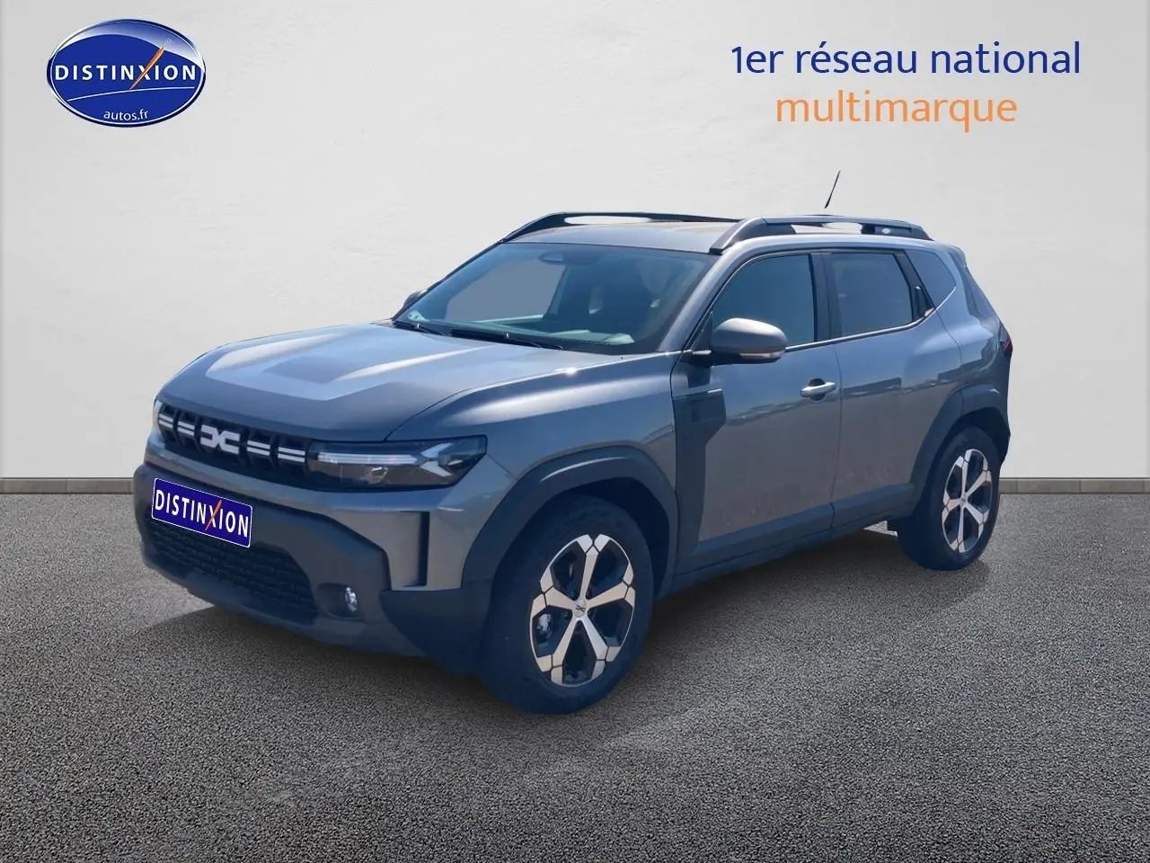 Dacia Duster gris schiste en 3/4 avant droit, avec jantes alliage et barres de toit visibles.