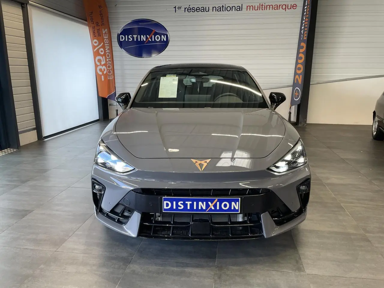 Vue avant d'une CUPRA Leon gris magnétique avec logo cuivré, phares allumés et calandre noire en showroom.