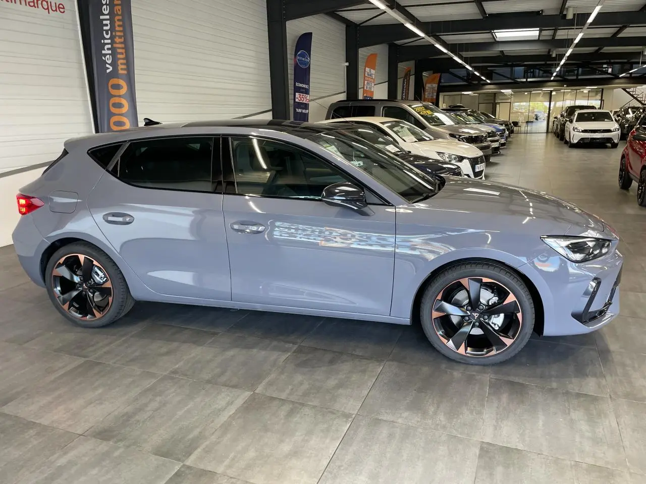 Vue de profil côté gauche d'une CUPRA Leon gris magnétique avec jantes alliage noires et cuivrées en showroom.