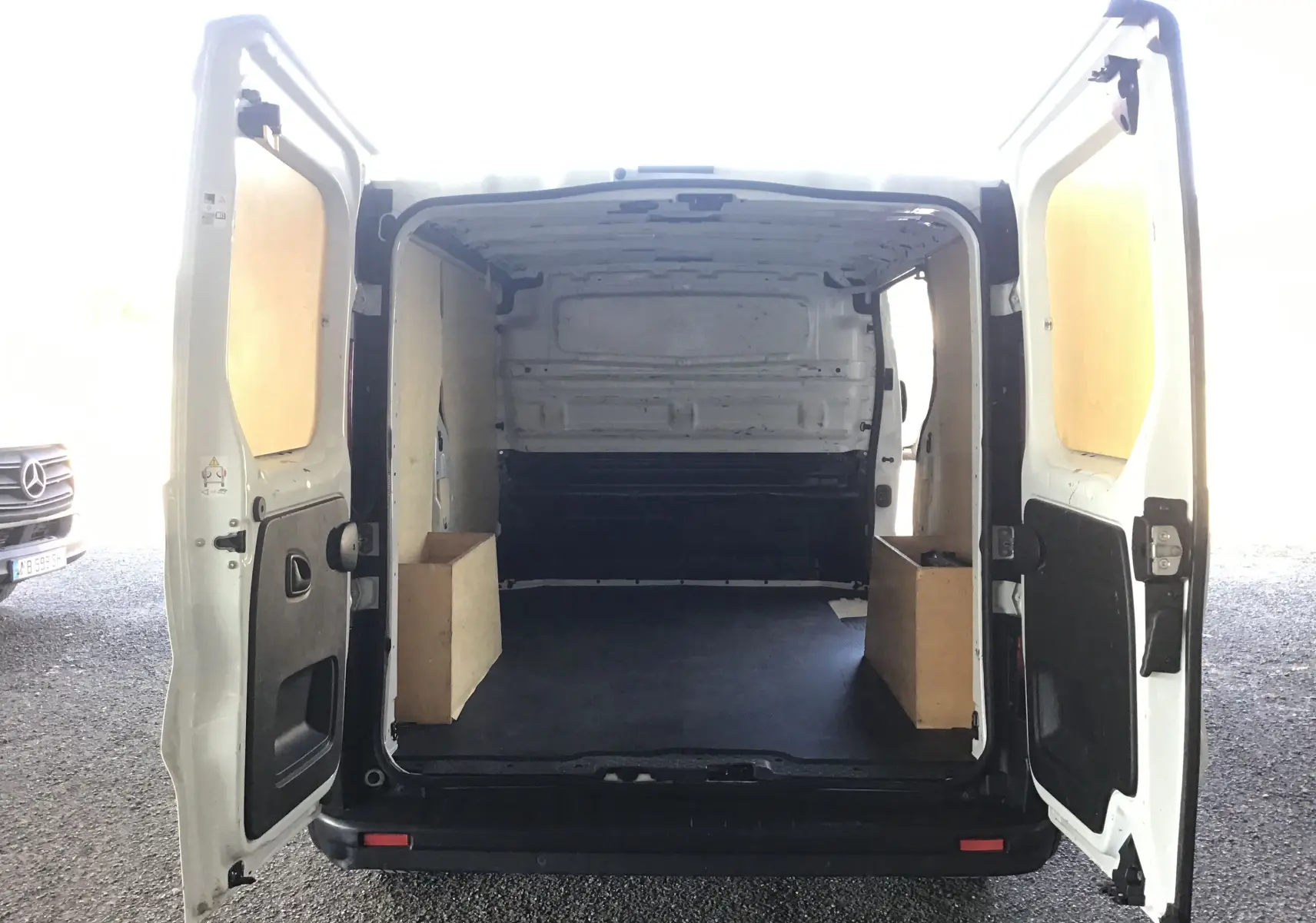 Vue arrière du fourgon blanc Nissan Primastar L1H1 2022 avec portes ouvertes montrant l'espace de chargement vide et doublé en bois.