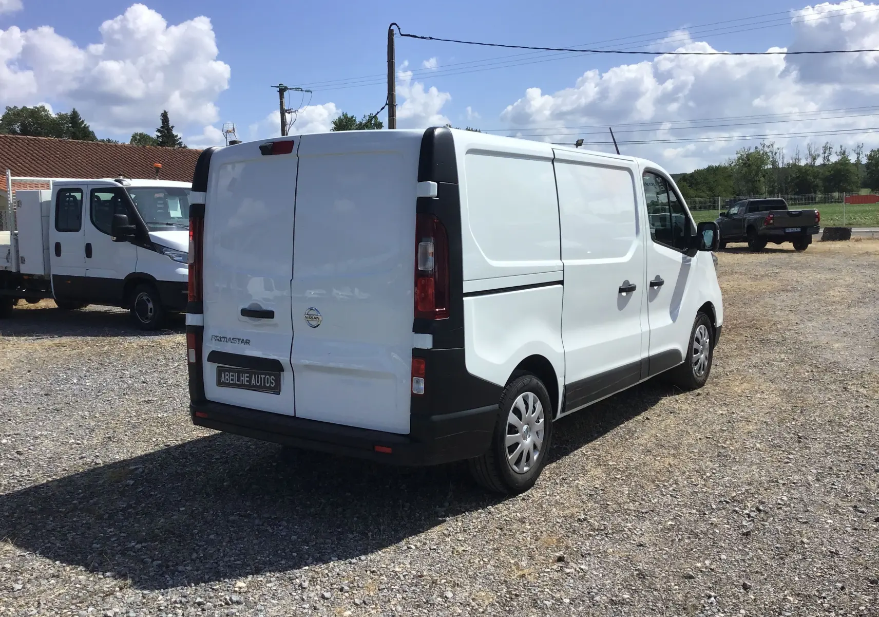 Fourgon blanc Nissan Primastar L1H1 vu en 3/4 arrière droit sur un parking ensoleillé avec ciel bleu.