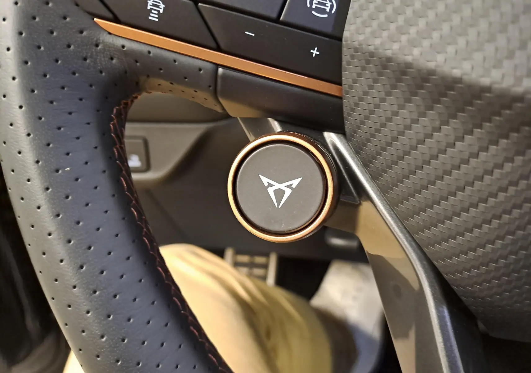 Gros plan sur le bouton rotatif du volant perforé avec logo CUPRA, entouré de finitions cuivre et carbone.