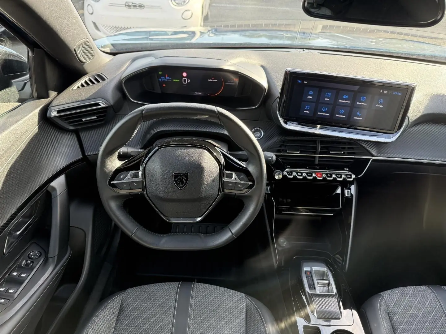 Vue intérieure avant du poste de conduite du Peugeot e-2008 2025 avec volant compact, écran tactile et finition carbone noire.