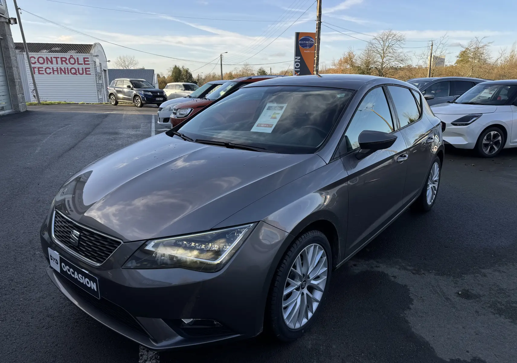 Vue 3/4 avant d'une SEAT Leon gris métallisé 2016 garée sur un parking avec phares allumés.