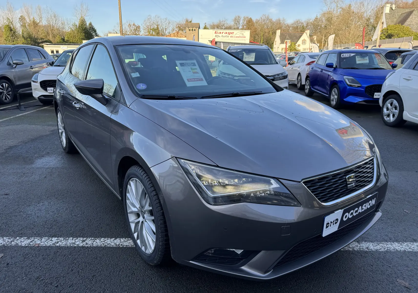 SEAT Leon gris vue 3/4 avant droit sur parking, avec phares allumés et jantes alliage visibles.