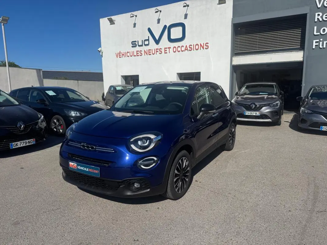Fiat 500X bleu vue 3/4 avant droit, avec phares ronds et jantes classiques 16 pouces, stationnée devant un garage.