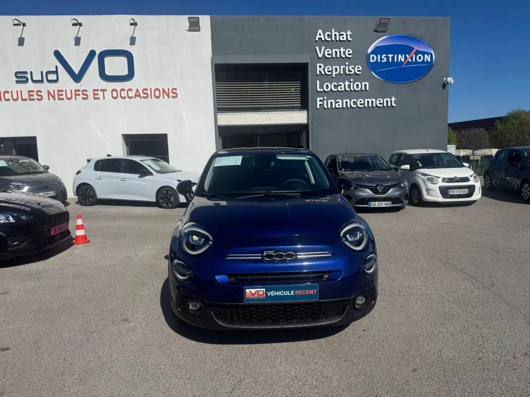 Vue de face d'une FIAT 500X bleu foncé stationnée devant un garage avec panneaux publicitaires visibles.