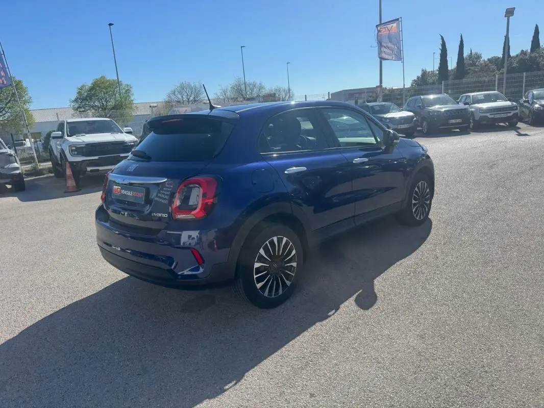 Fiat 500X bleu en 3/4 arrière droit sur parking, avec jantes classiques 16 pouces et feux arrière distinctifs.