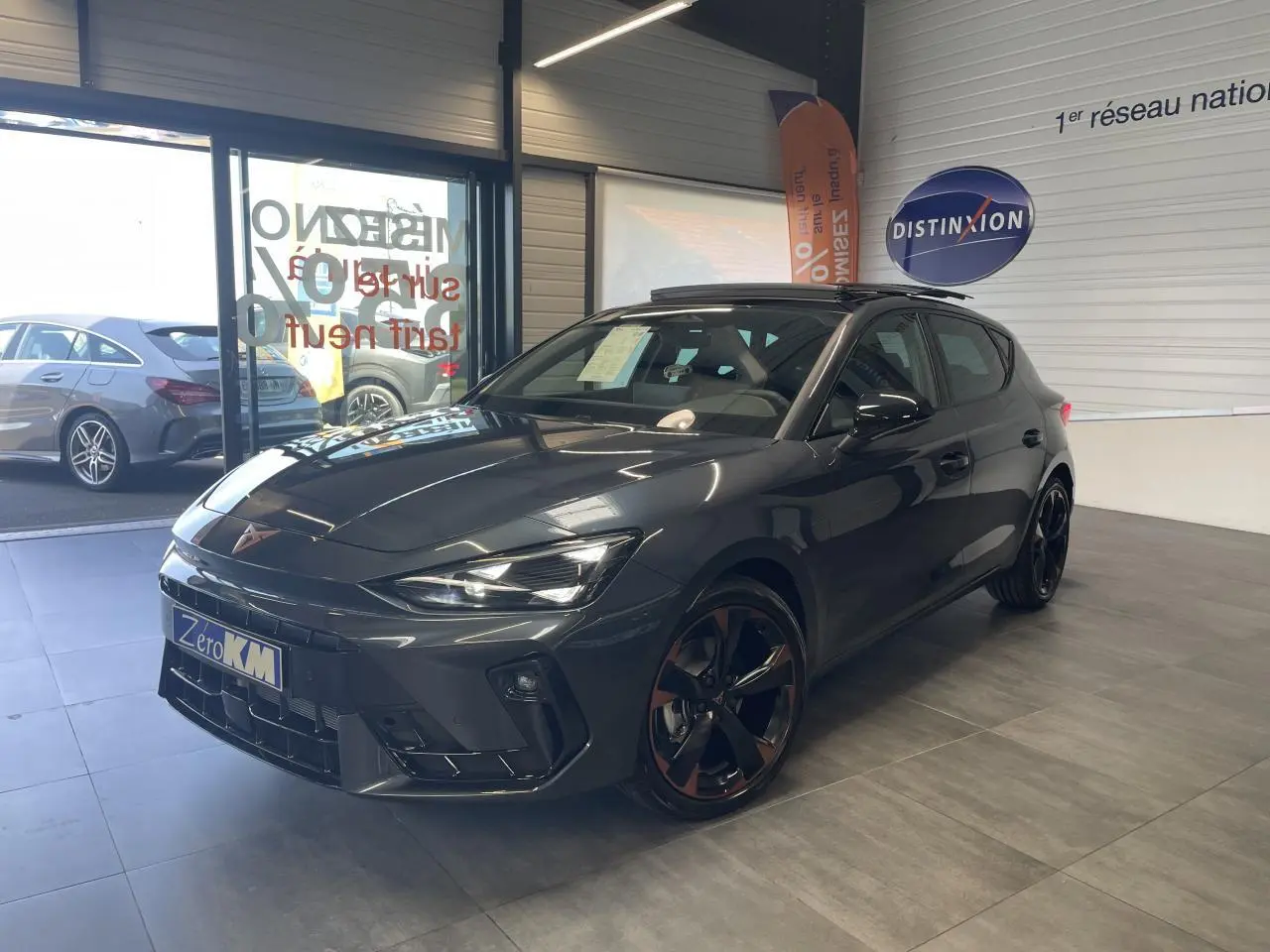 Vue 3/4 avant d'une CUPRA Leon gris magnétique avec jantes cuivrées et toit ouvrant panoramique en showroom.