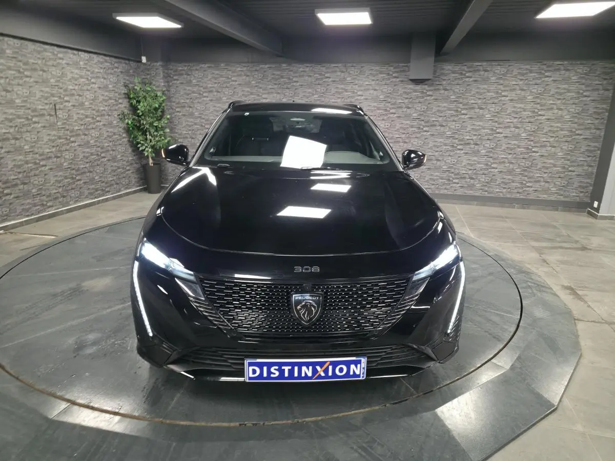 Vue avant d'une Peugeot 308 SW GT noire 2024 avec calandre noire brillante et feux LED allumés dans un showroom.