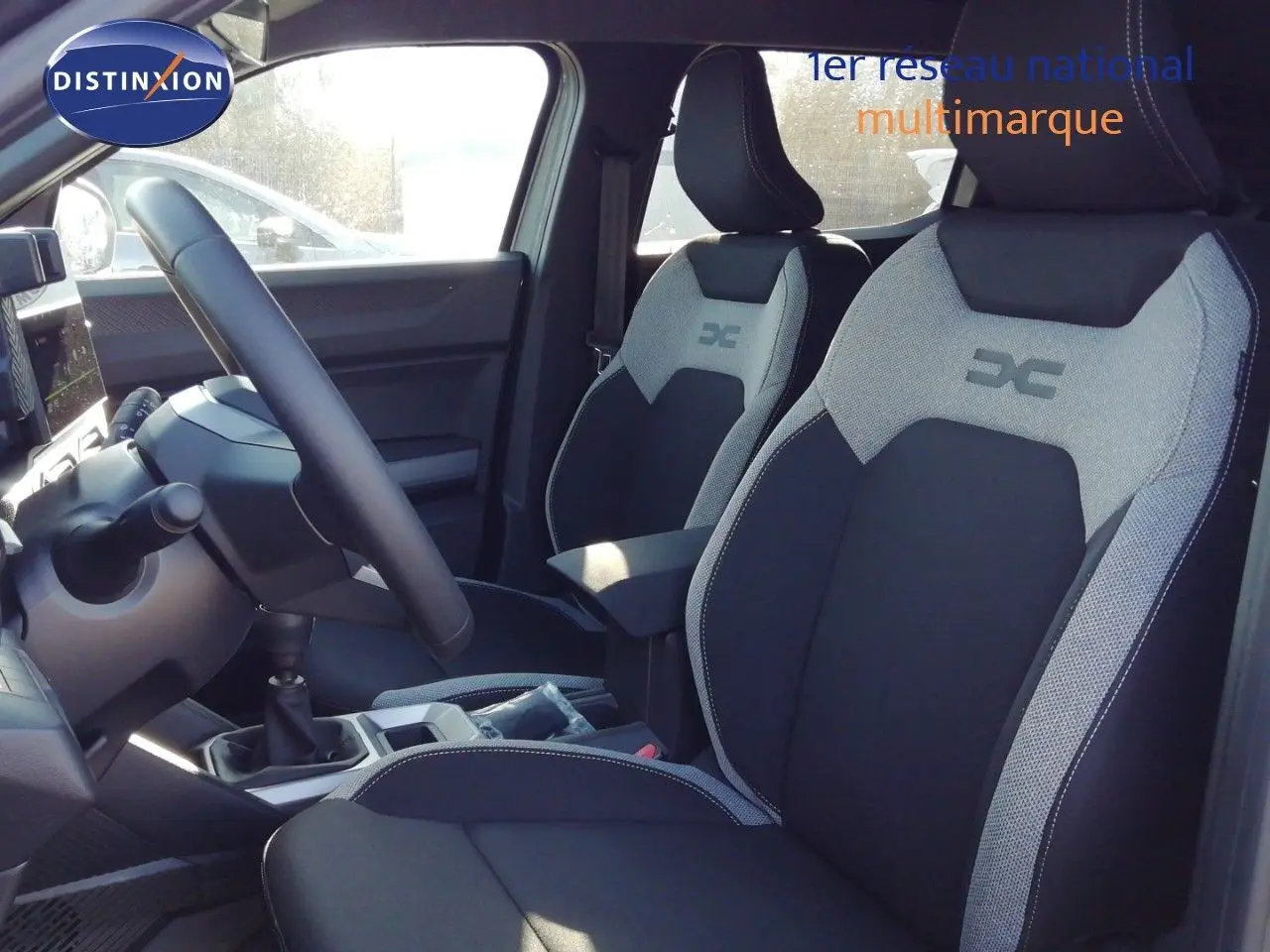 Vue intérieure côté conducteur du Dacia Duster 2026, montrant les sièges bi-ton gris et noir avec logo DC brodé.