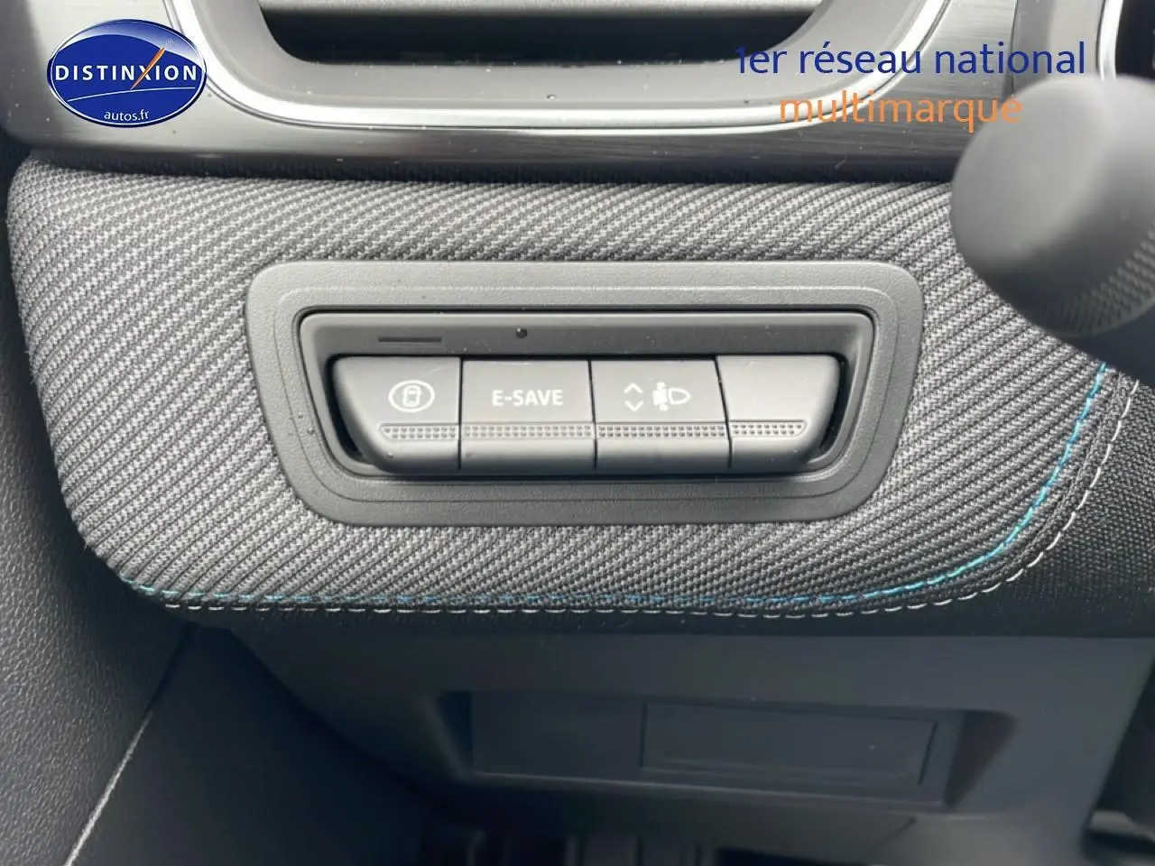 Gros plan sur les commandes E-Save et réglage des phares sur tableau de bord en tissu gris avec surpiqûres bleues.
