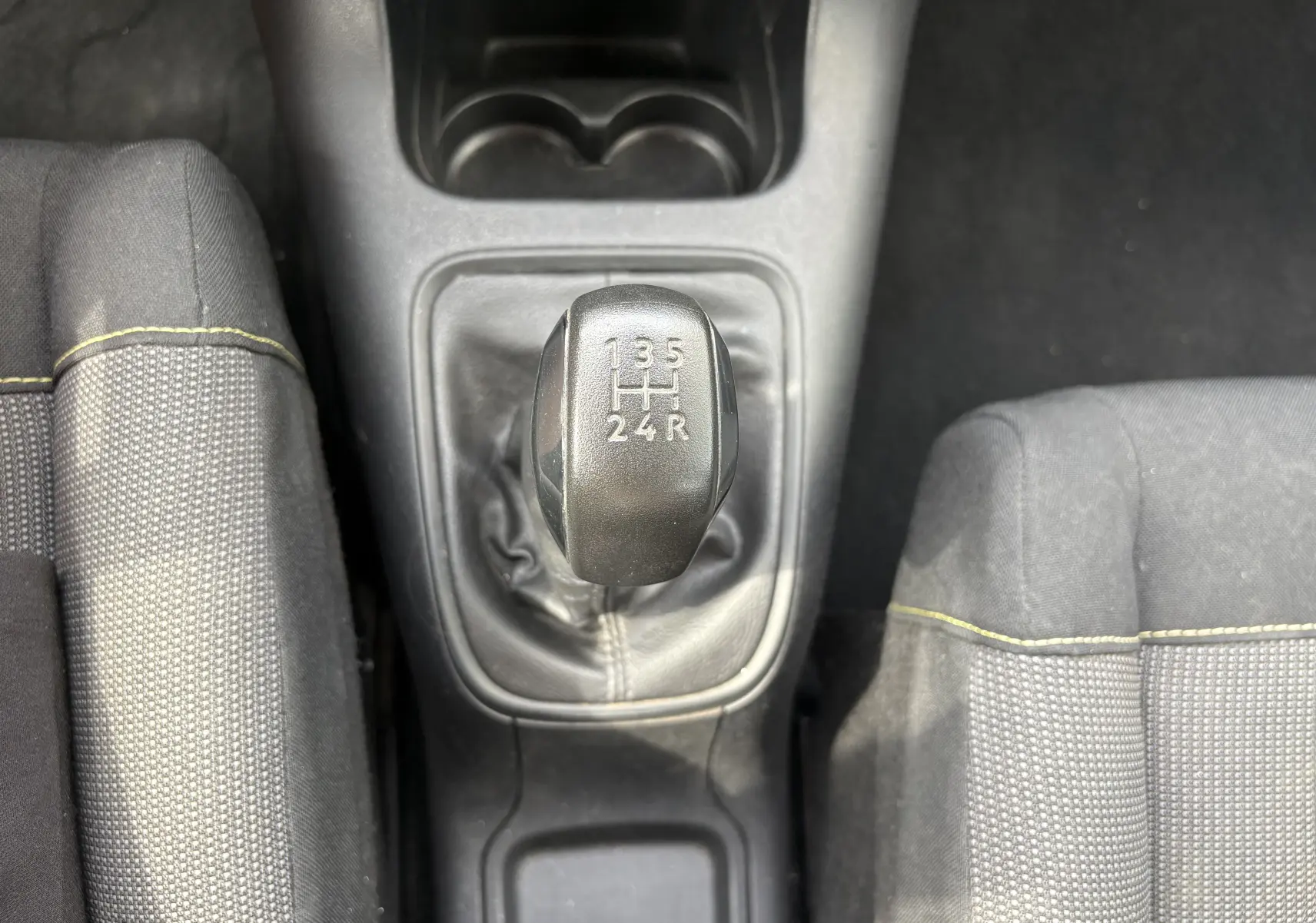 Vue plongeante sur le levier de vitesses manuel noir d’une Citroën C3 blanche, entouré des sièges gris à surpiqûres jaunes.