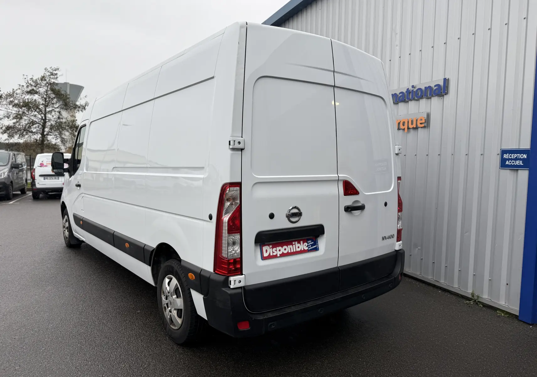 Vue 3/4 arrière droite d'un utilitaire Nissan NV400 blanc, fourgon long avec portes arrière fermées sur parking humide.