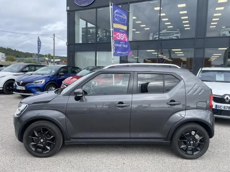 Profil côté gauche d'une Suzuki Ignis 1.2 Dualjet Hybrid gris foncé métal avec jantes noires, vue en extérieur devant un showroom.