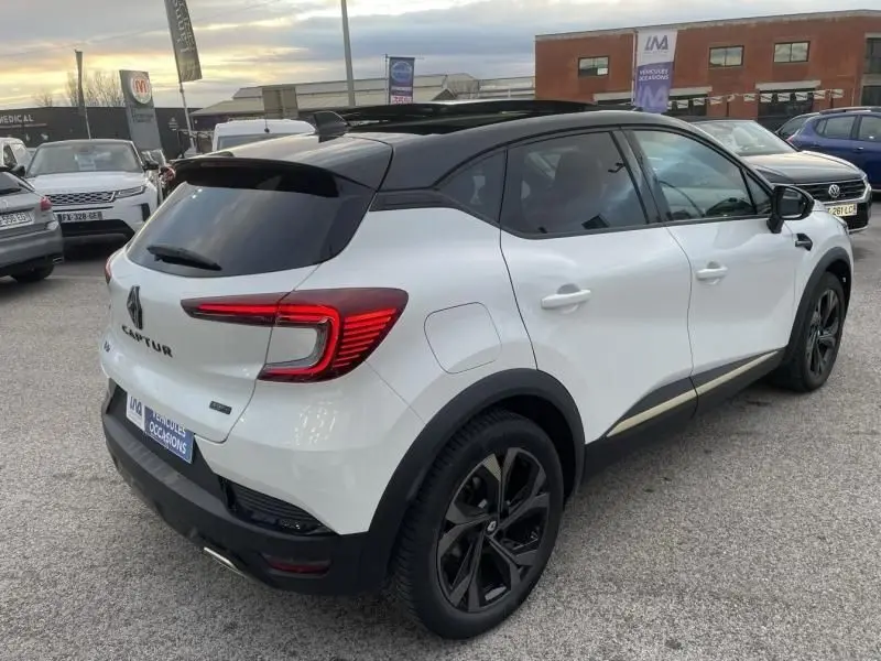 Vue 3/4 arrière droite d’un Renault Captur blanc nacré 2023 avec toit noir et jantes noires, version hybride E-Tech Engineered.