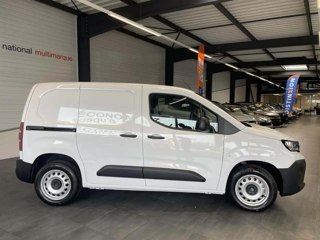Vue de profil côté droit d'un Peugeot Partner blanc avec jantes tôles grises en intérieur de showroom.