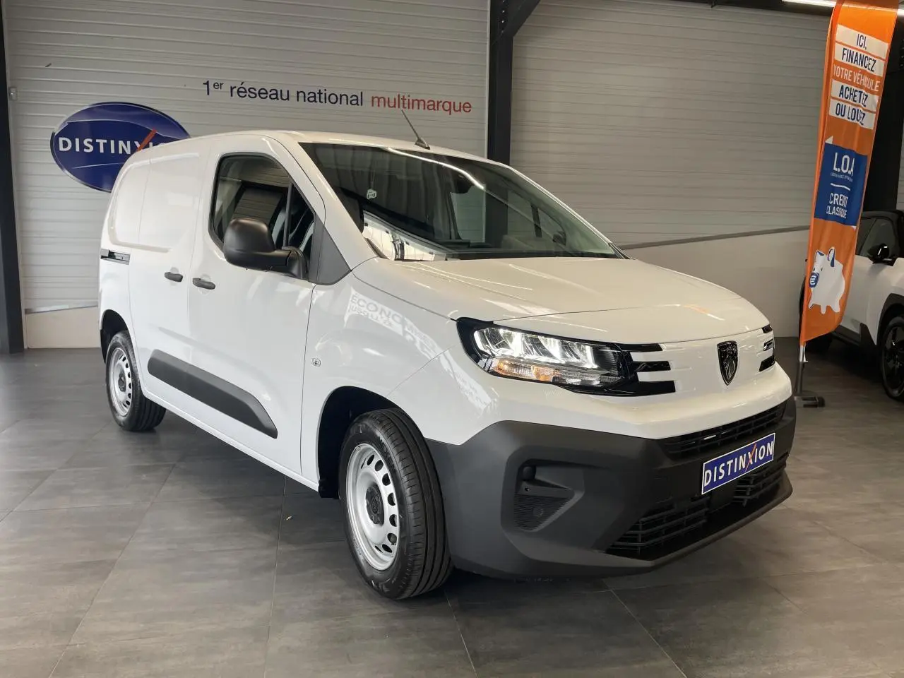 Vue 3/4 avant droit du fourgon blanc Peugeot Partner 2026 avec pare-chocs noirs et jantes tôles grises en showroom.