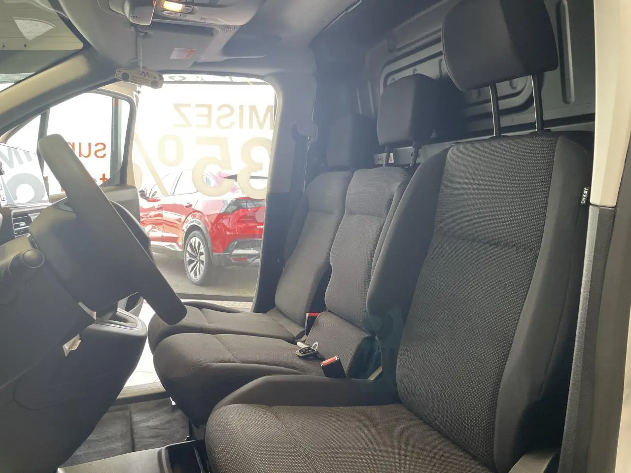 Vue intérieure côté conducteur du Peugeot Partner blanc, montrant la banquette passager 3 places en tissu noir.