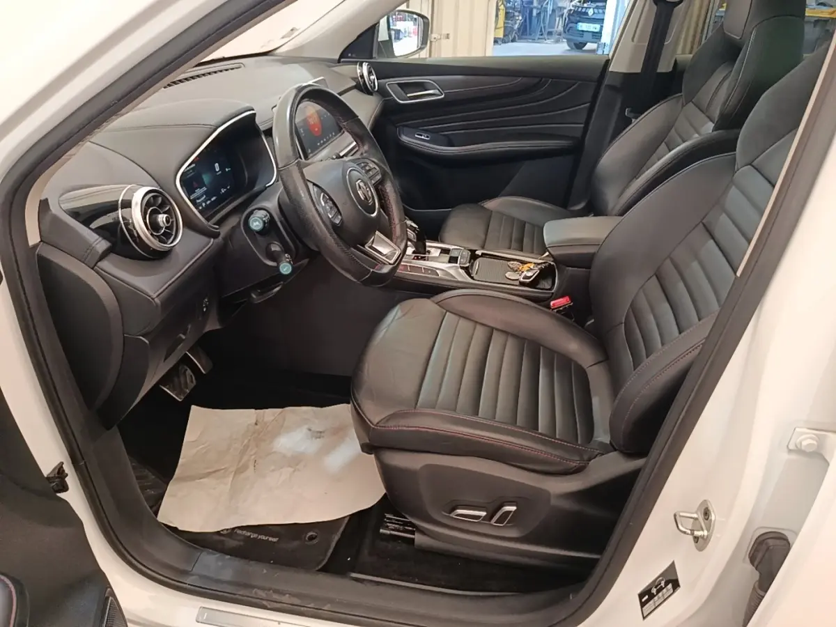 Vue intérieure côté conducteur du MG Motor EHS blanc 2023, sièges cuir noirs avec surpiqûres rouges et tableau de bord digital.