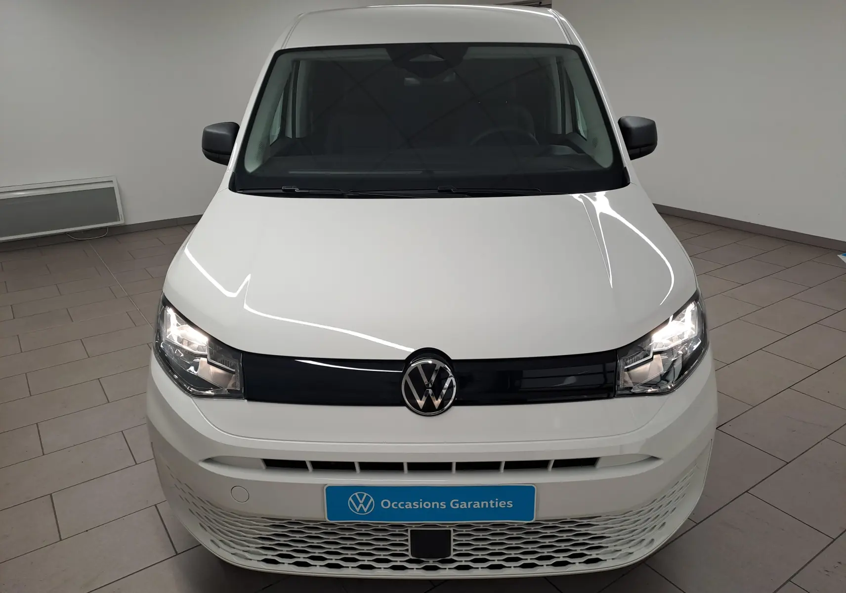 Vue frontale d’un Volkswagen Caddy Cargo Maxi blanc candy 2026 avec calandre noire et phares allumés en intérieur showroom.