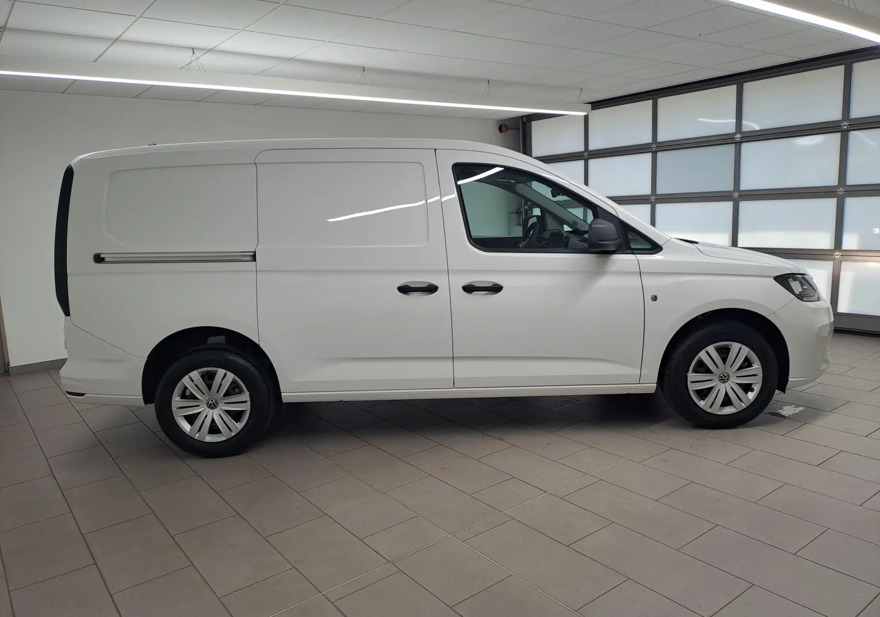 Vue de profil côté droit d'un Volkswagen Caddy Cargo Maxi blanc Candy 2026 en intérieur, avec portes latérales coulissantes.