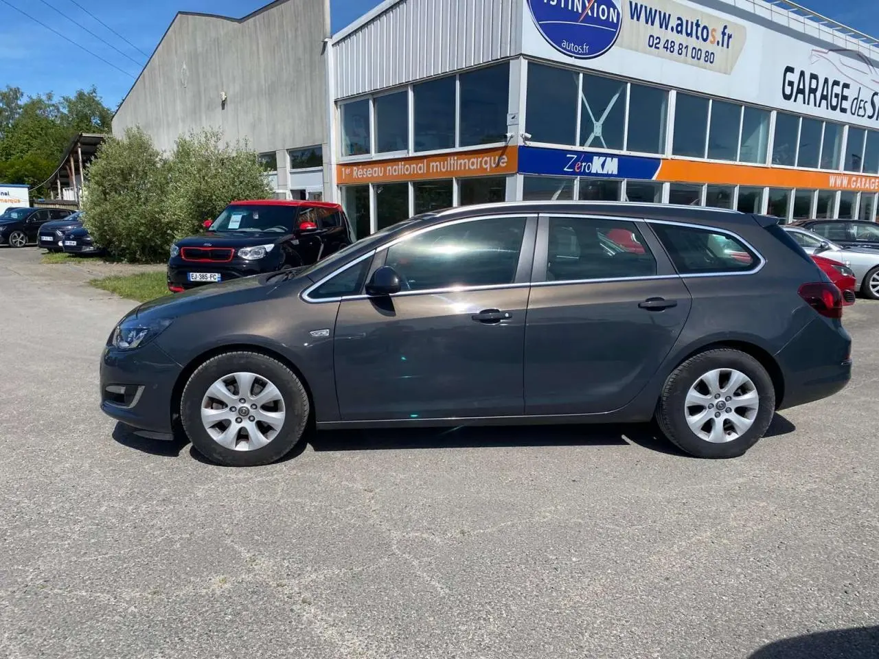 Opel Astra Sports Tourer gris vue de profil côté gauche, garée devant un garage multimarque.