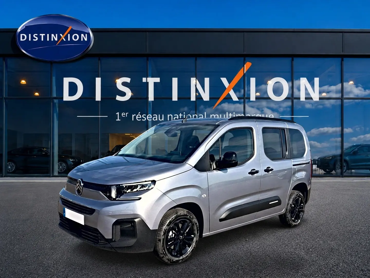 Citroën Berlingo Steel Grey en 3/4 avant droit, avec jantes noires et barres de toit visibles devant un bâtiment Distinxion.