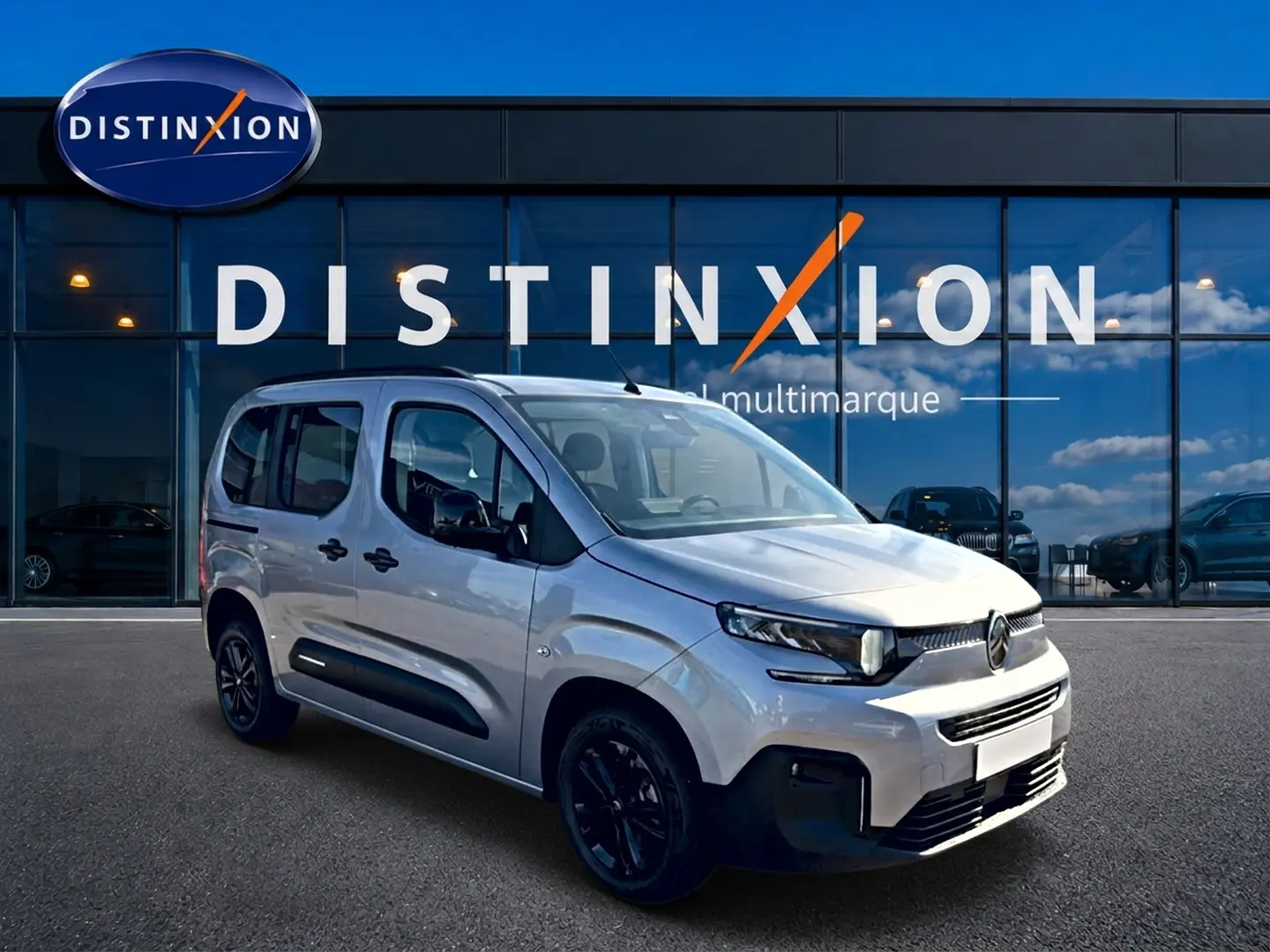 Vue 3/4 avant droit du Citroën Berlingo 2026 gris acier avec barres de toit et jantes noires devant un showroom Distinxion.