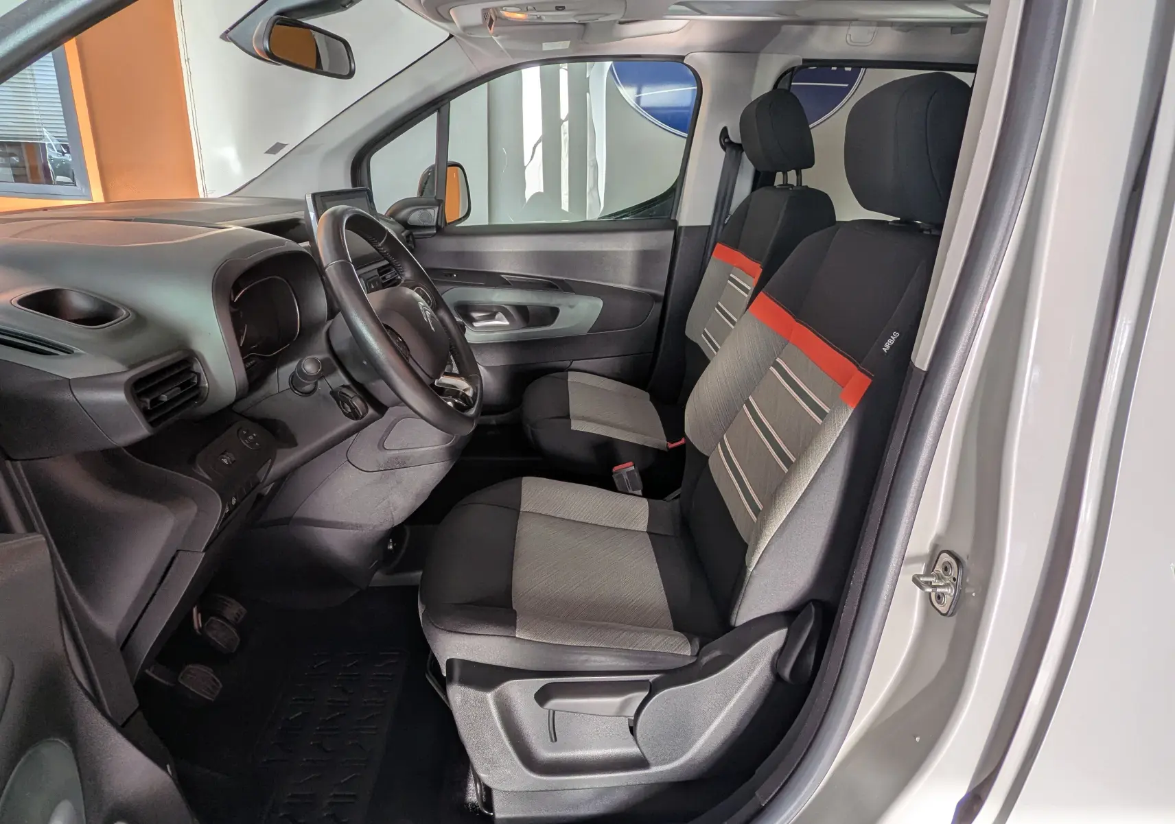 Vue intérieure côté conducteur du Citroën Berlingo beige 2019, sièges tissu gris avec bande orange et tableau de bord noir.
