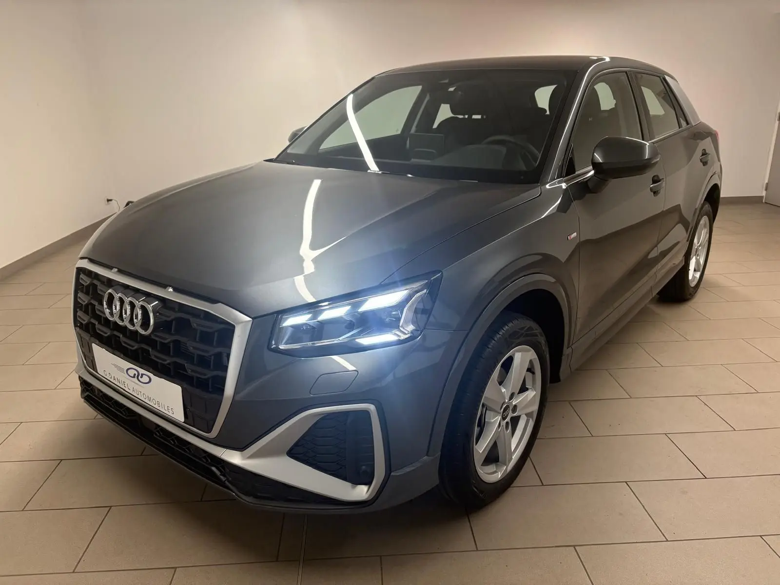 Audi Q2 35 TFSI S Line gris Daytona en 3/4 avant droit avec phares allumés dans un intérieur neutre.