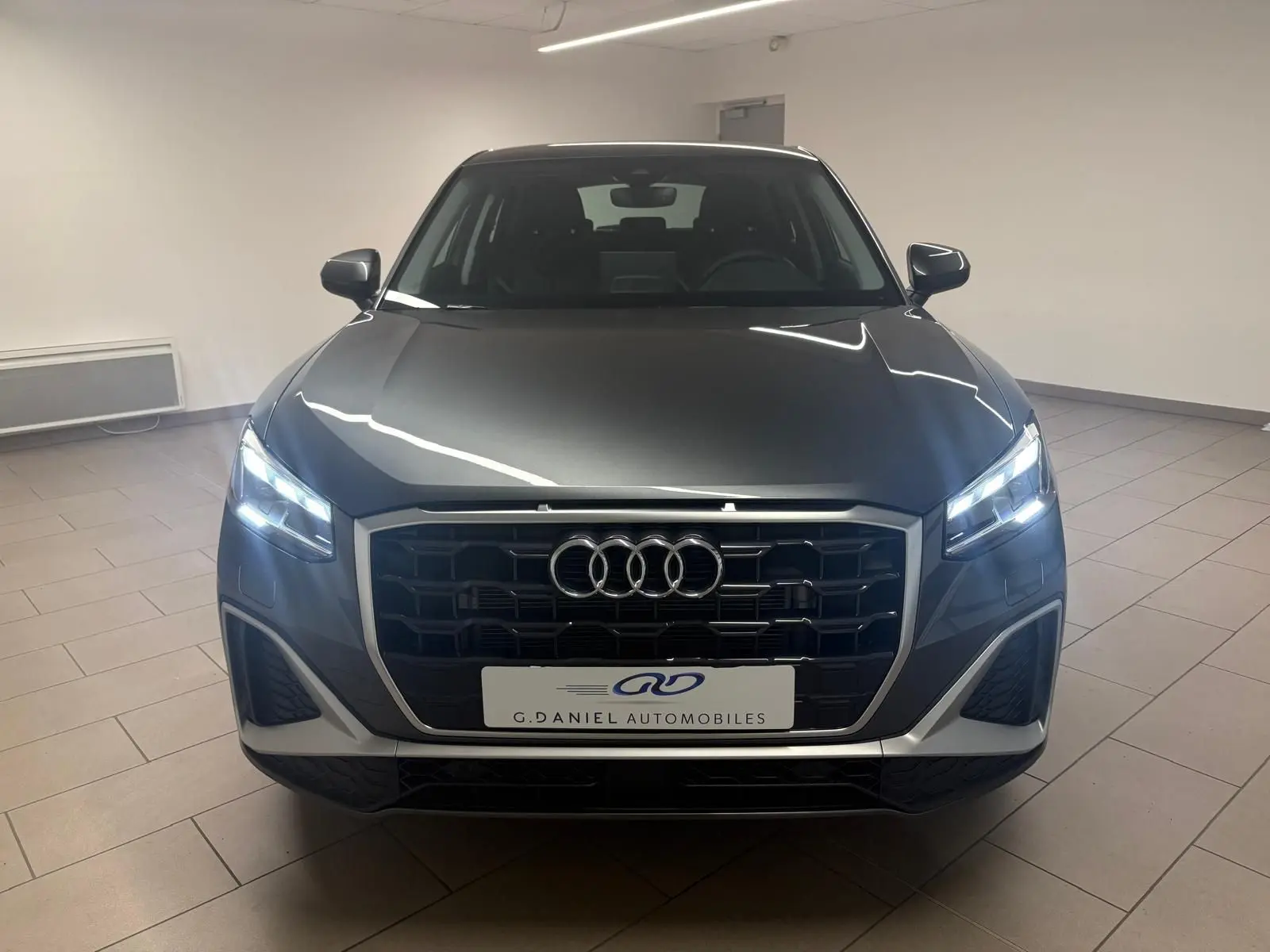 Vue avant d'un Audi Q2 35 TFSI S Line gris Daytona avec phares allumés dans un intérieur neutre.