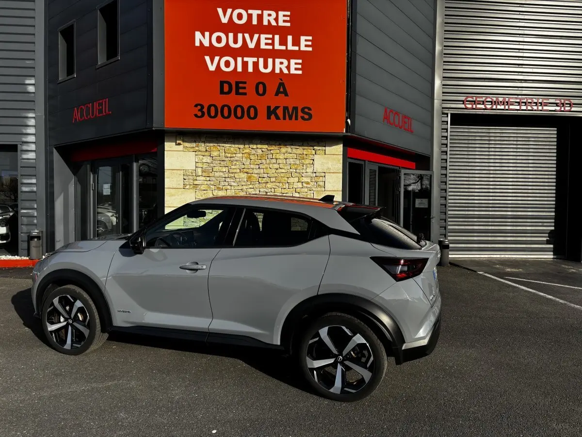 Vue de profil côté gauche d’un Nissan Juke Hybrid gris clair 2024 garé devant un bâtiment avec panneau orange.