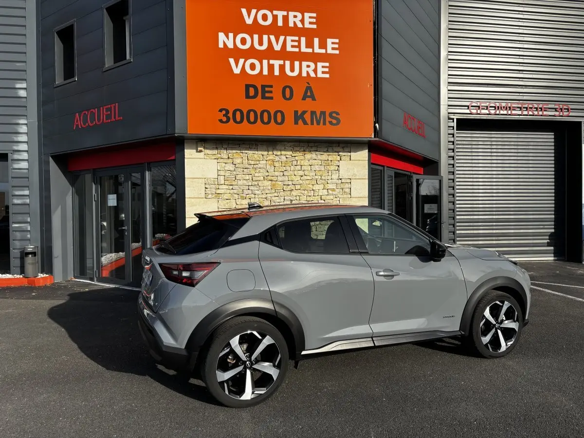 Vue de profil droit d'un Nissan Juke Hybrid gris clair avec toit noir, garé devant un bâtiment commercial.