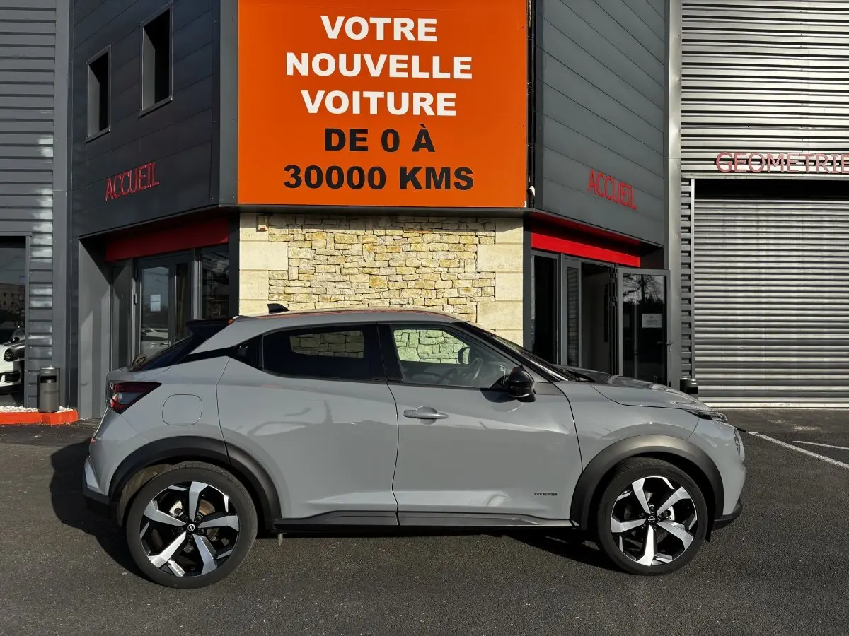 Profil droit d'un Nissan Juke Hybrid gris clair 2024 avec jantes alliage noires et toit noir, stationné devant un bâtiment.