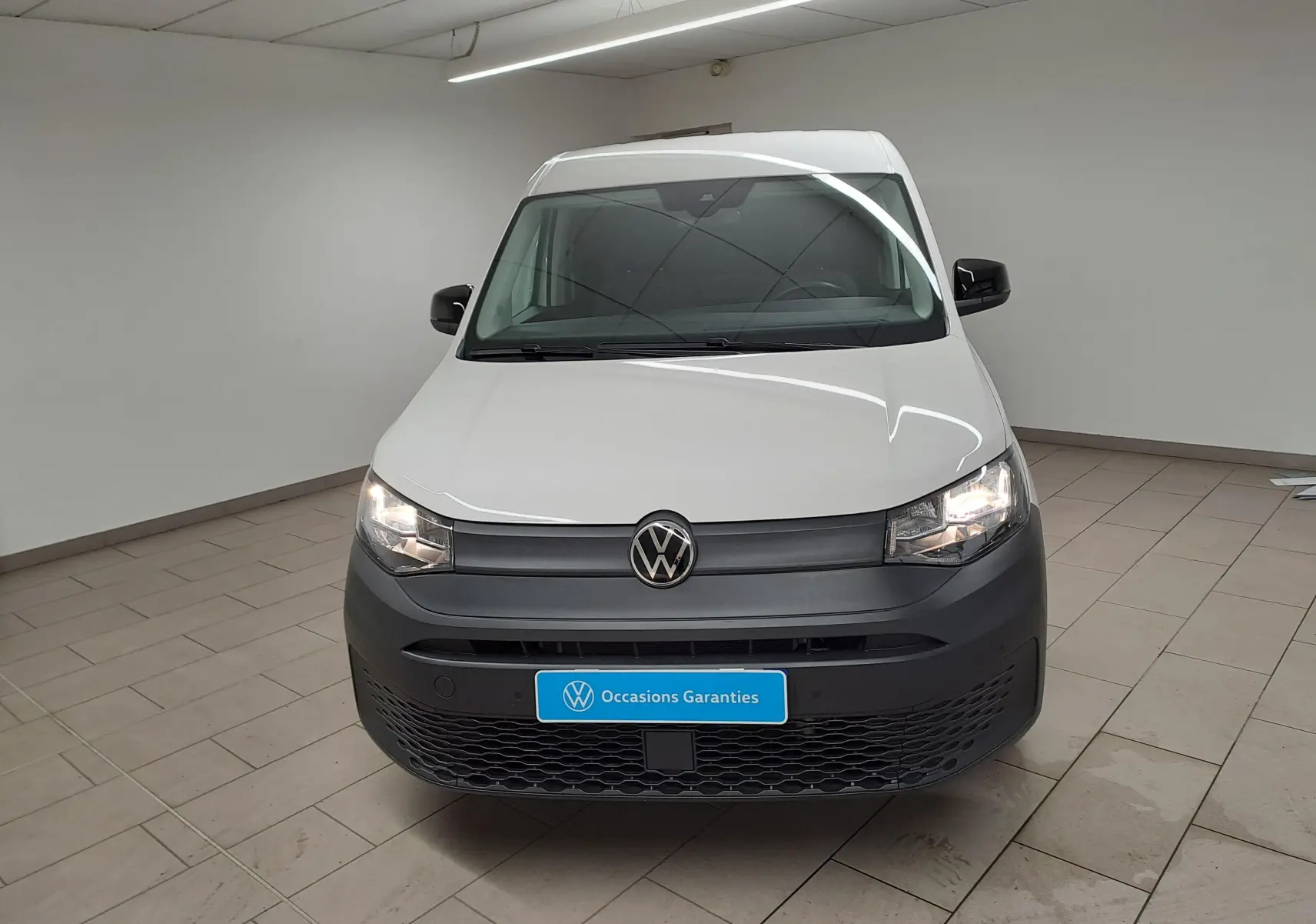Vue avant d'un Volkswagen Caddy blanc Candy 2025 avec pare-chocs noir mat et phares allumés en intérieur.