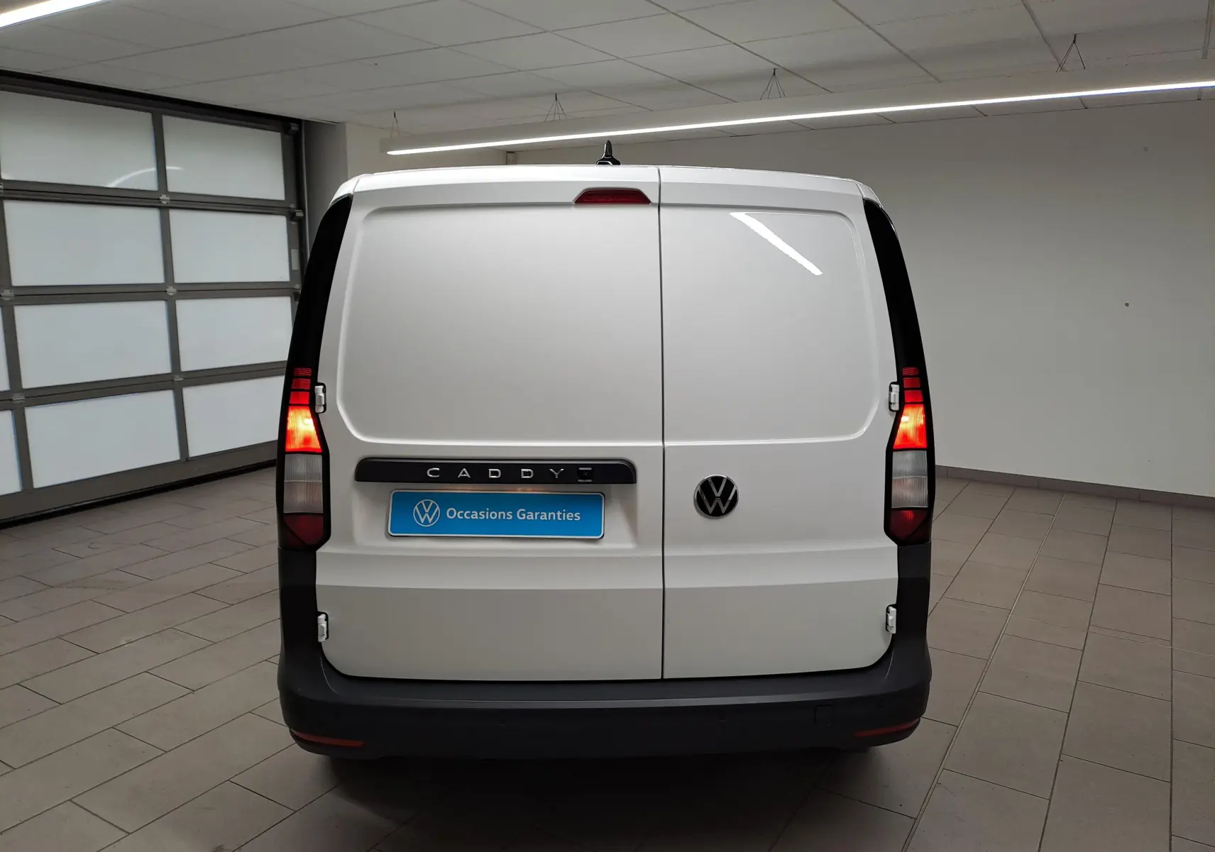 Vue arrière d'un Volkswagen Caddy blanc Candy avec feux arrière allumés et logo VW visible.