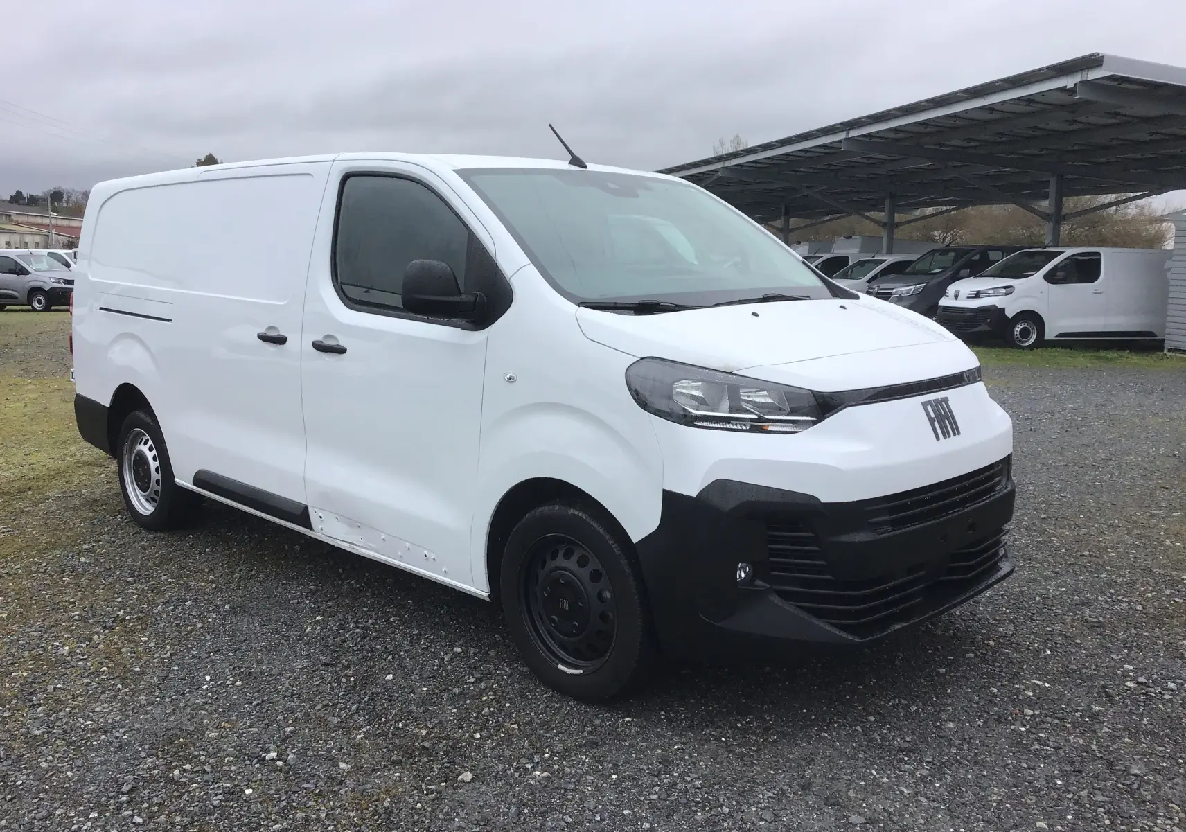 Vue 3/4 avant droite d’un FIAT Scudo Fourgon blanc 2024 avec pare-chocs noir et jantes tôles noires, stationné sur gravier.