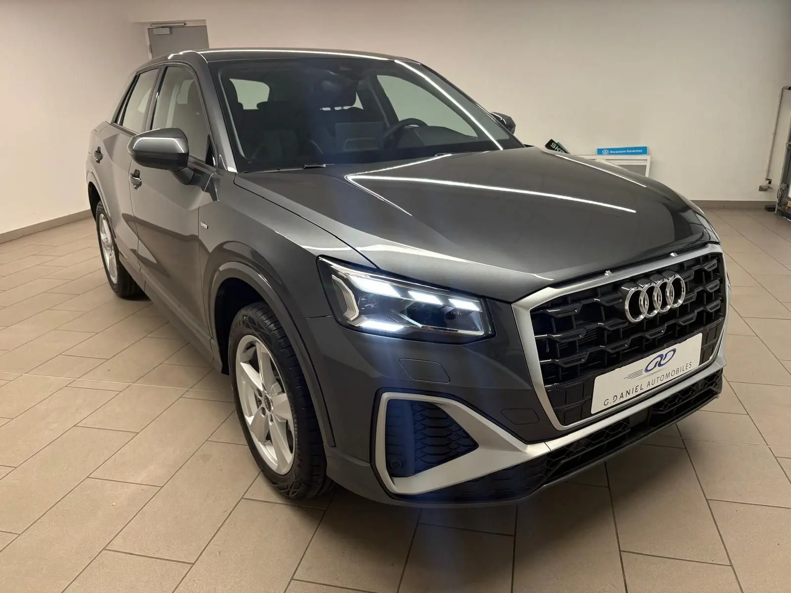 Audi Q2 35 TFSI S Line gris Daytona en 3/4 avant droit, avec calandre noire et phares LED allumés.