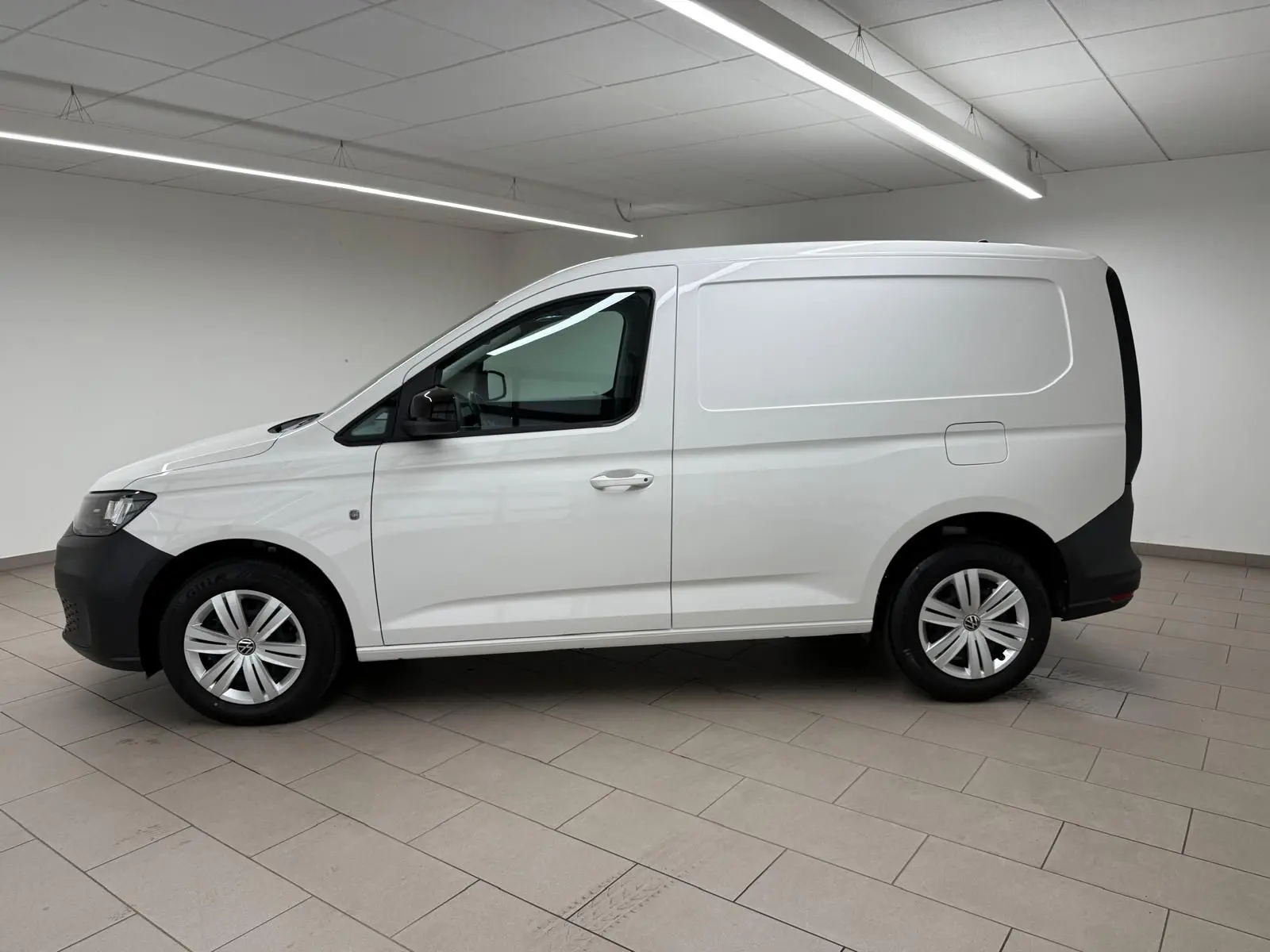 Vue de profil côté gauche d'un Volkswagen Caddy blanc Candy avec pare-chocs et protections noires, en intérieur.