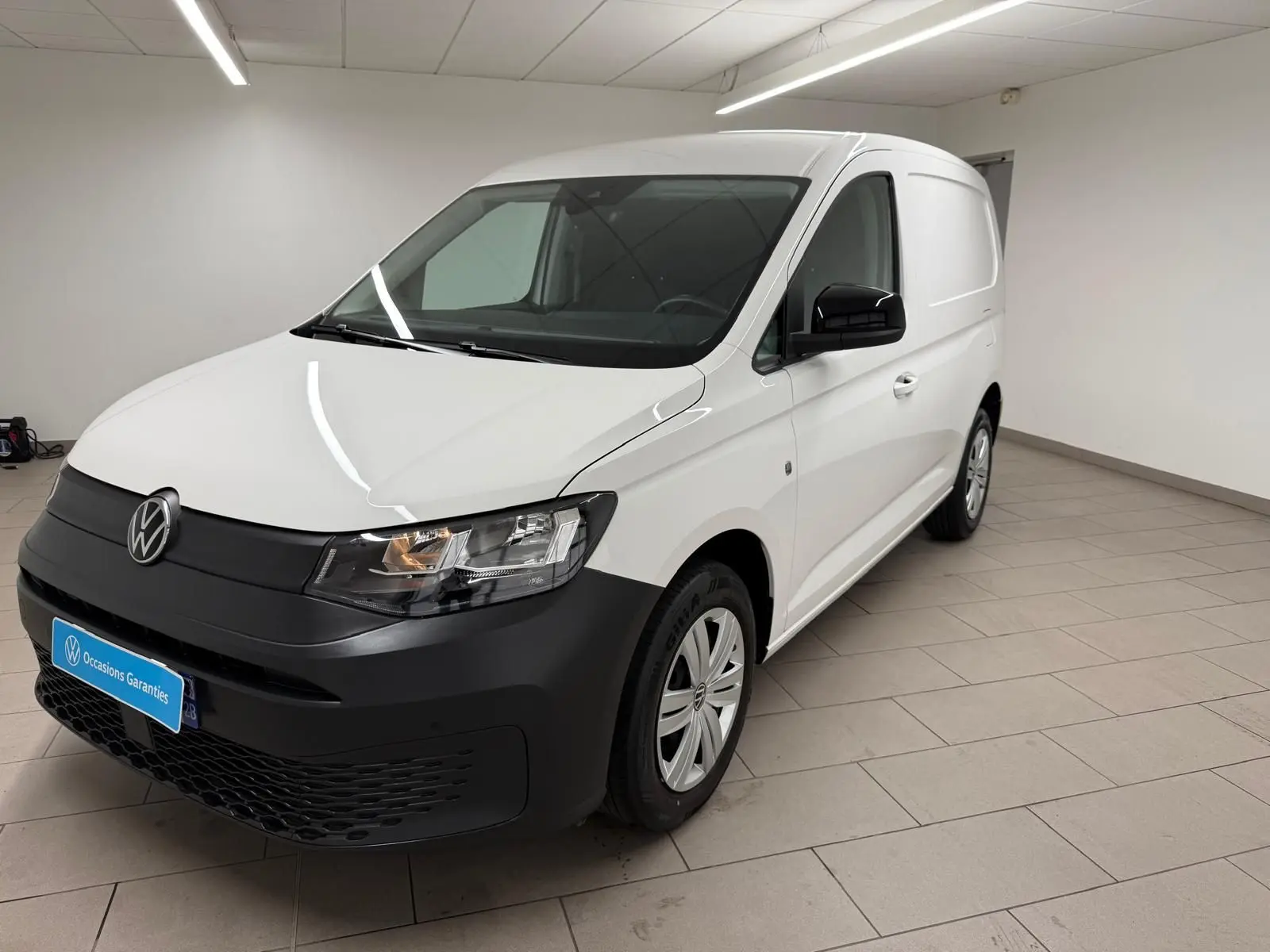 Vue 3/4 avant droit du Volkswagen Caddy blanc Candy avec pare-chocs noir mat et jantes acier en intérieur showroom.