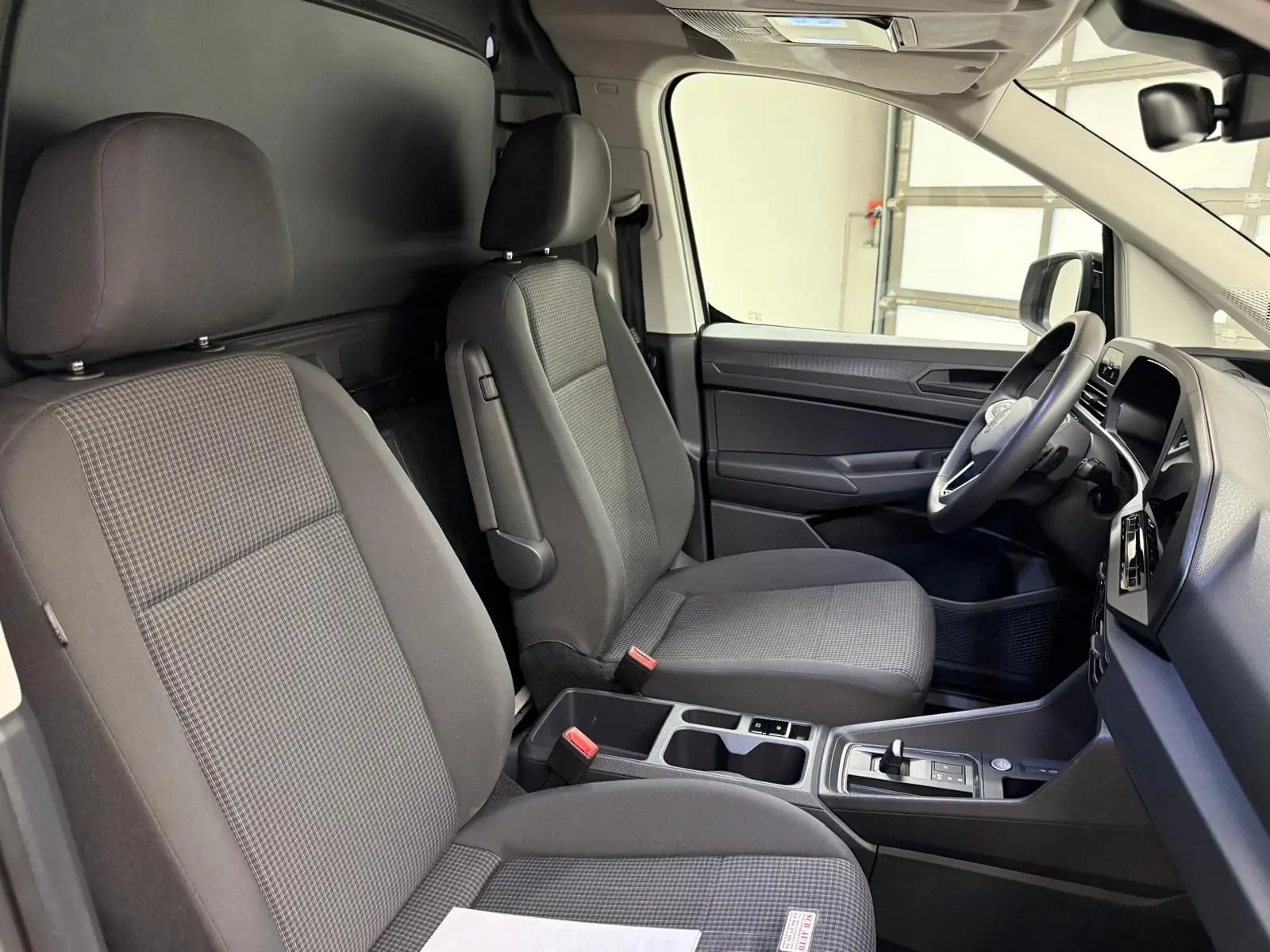 Vue intérieure côté conducteur du Volkswagen Caddy 2025, sièges tissu gris et console centrale avec levier de vitesses.