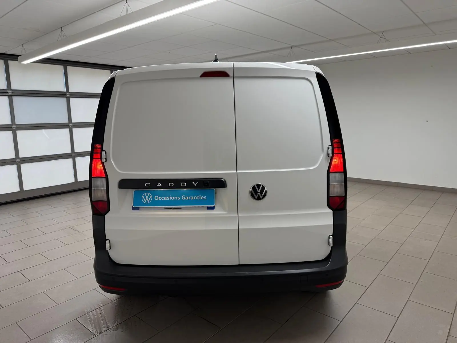 Vue arrière d'un Volkswagen Caddy blanc Candy avec portes battantes et feux arrière allumés en intérieur.
