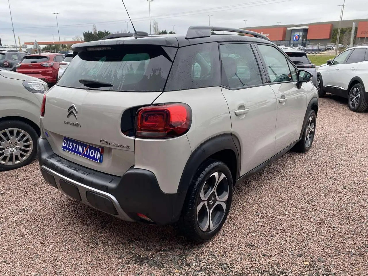Vue 3/4 arrière droite d’un Citroën C3 Aircross gris 2020 avec toit noir et jantes distinctives, stationné sur un sol gravillonné.