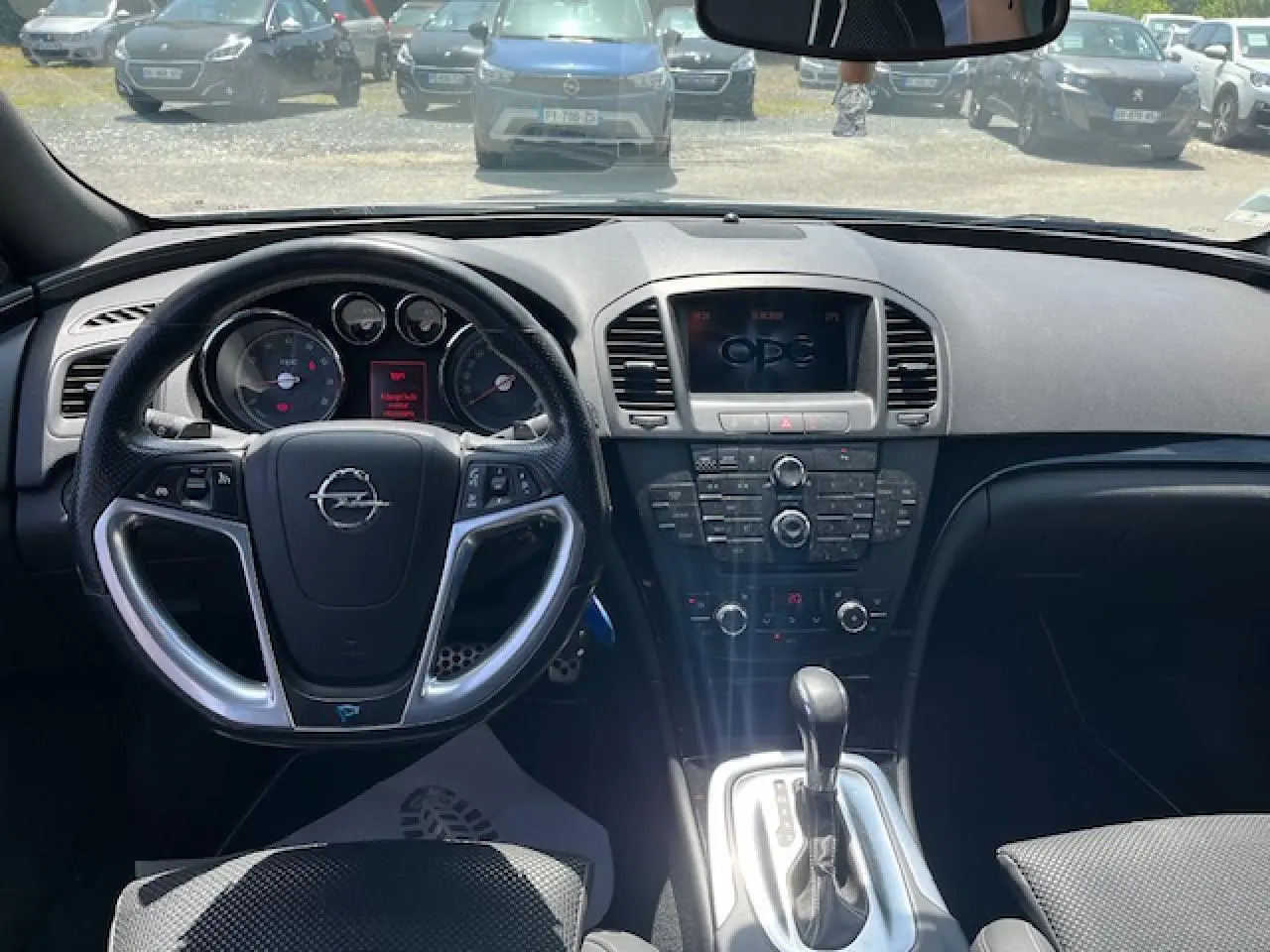 Vue intérieure centrée sur le tableau de bord et volant d’une Opel Insignia Sports Tourer gris clair, avec console centrale et levier de vitesse automatique.