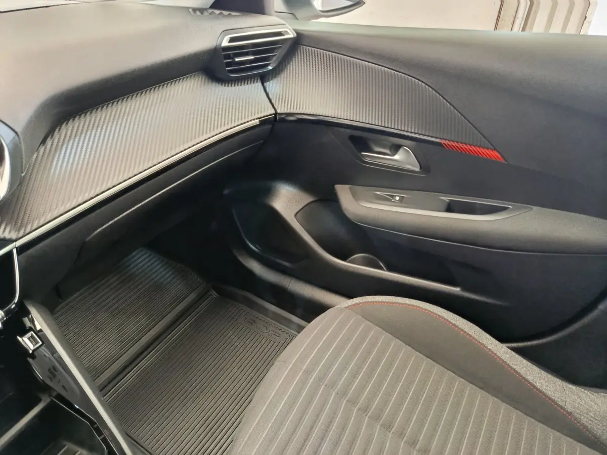 Intérieur côté passager de la Peugeot 208 PureTech 75 blanc, avec tableau de bord noir texturé et siège tissu gris à surpiqûres rouges.